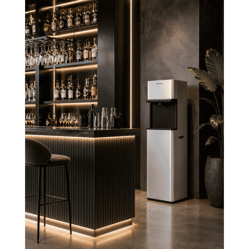 A tall stainless steel water dispenser with a black top section stands in a modern bar or lounge setting with dark wood shelving, illuminated bottles, a stool, and a potted plant.