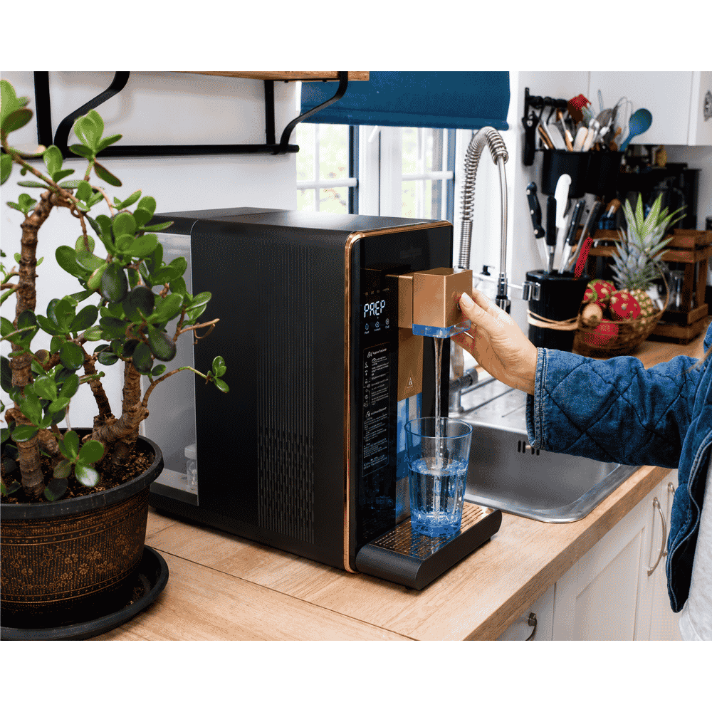 Person using a hydrogen water dispenser in a kitchen with a plant and utensils in the background.